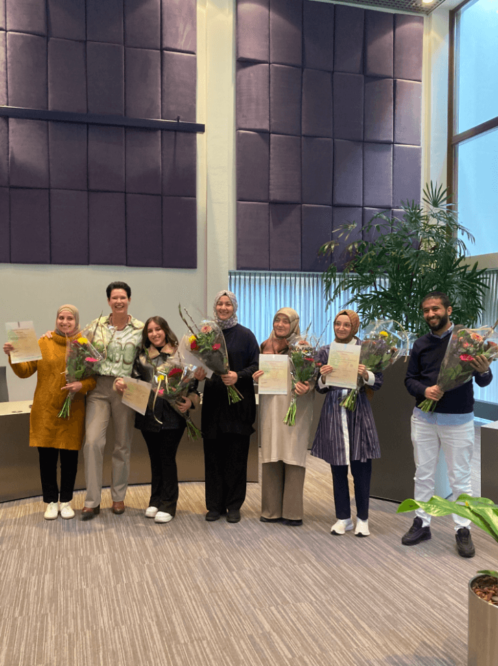 6 personen die bloemen en diploma's vasthouden en de wethouder
